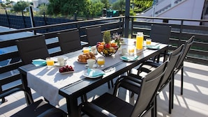A breakfast table set with croissants, fruit, juice, and a bowl of fruit on a terrace with chairs and a view of the surroundings. - Villa Residence Sunrise (Privlaka)