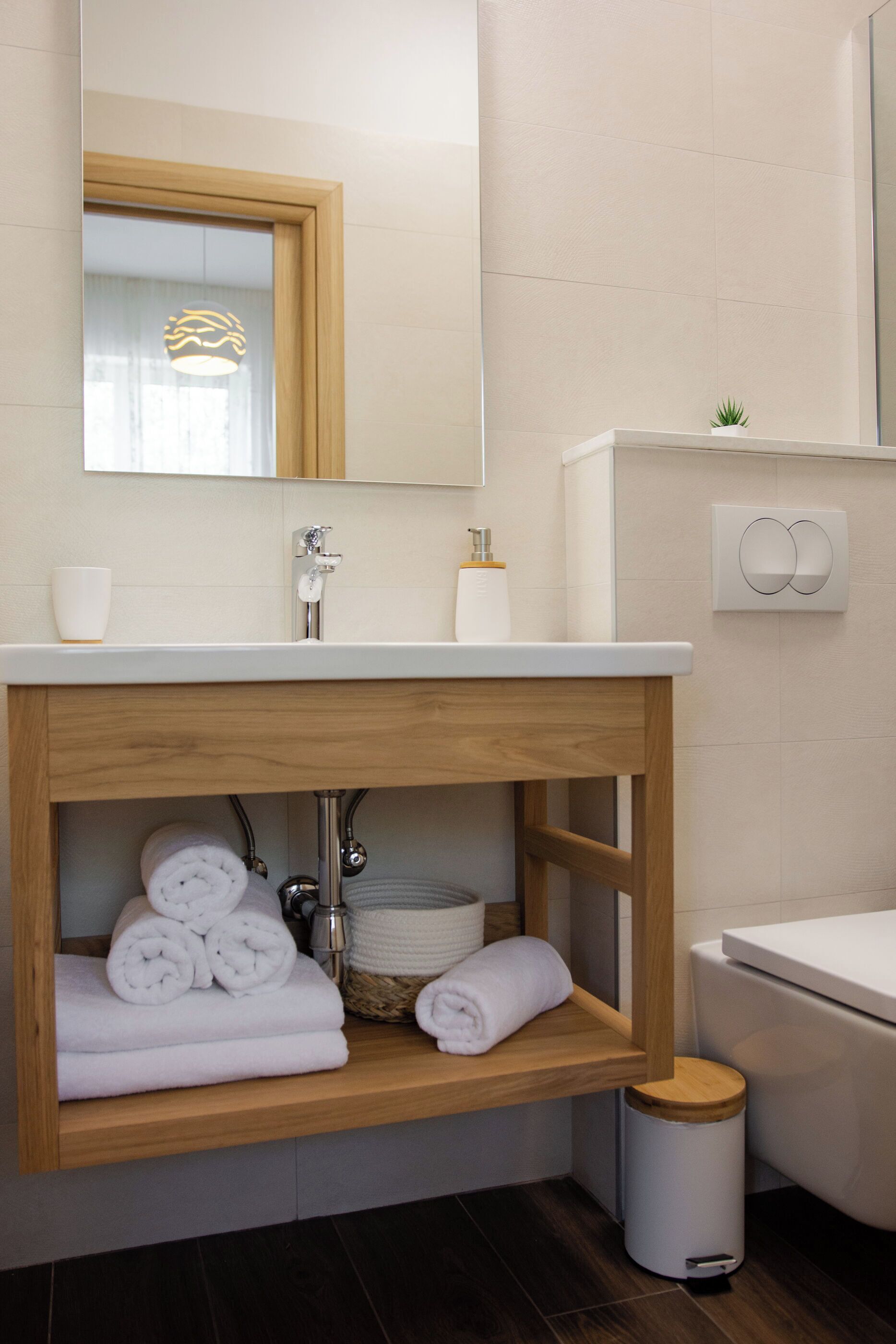 Ein modernes Badezimmer mit einem Holz-Waschtisch, einem weißen Waschbecken und einem Spiegel darüber. Auf dem Waschtisch liegen Handtücher und Pflegeprodukte, an der Wand steht eine kleine Pflanze.