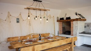 A wooden dining table set with plates, glasses, and a bottle, with a brick oven and hanging lights in the background. - Villa Residence Sunrise (Privlaka)