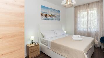 A neatly arranged bedroom with a bed, bedside table, lamp, and a painting of a lighthouse.