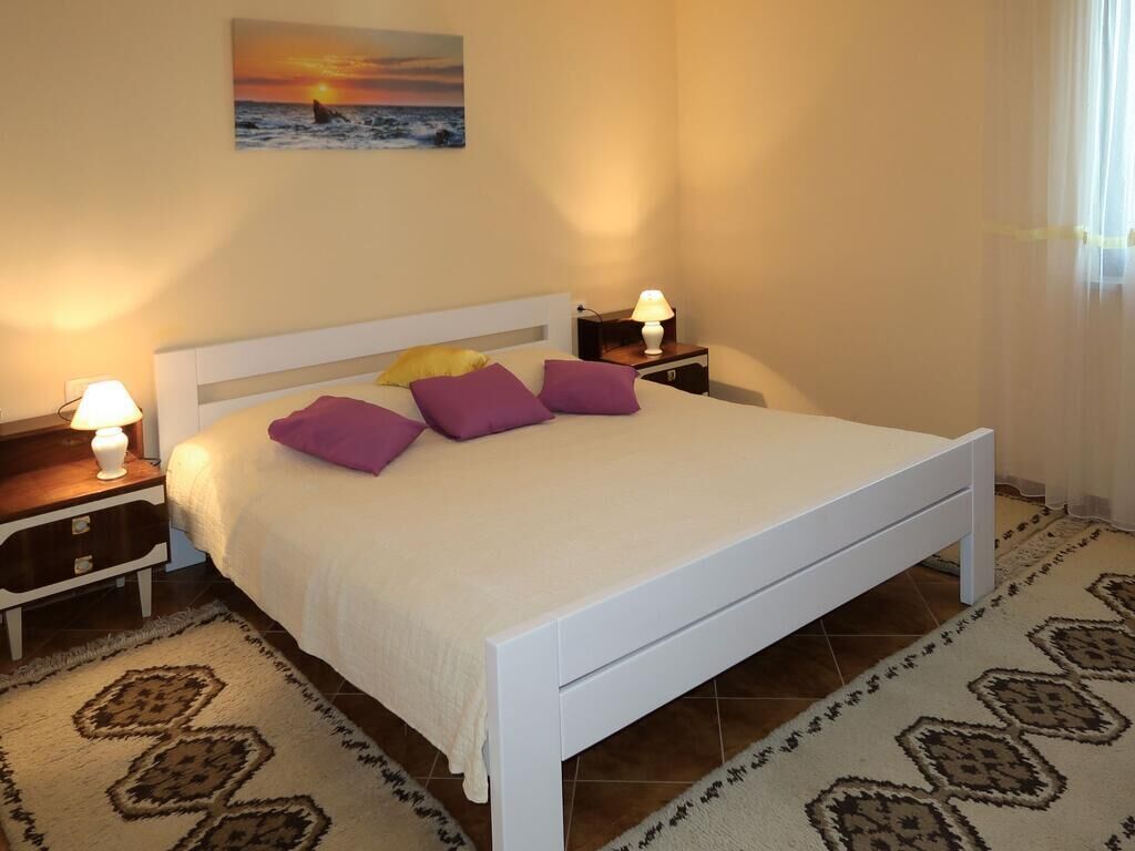 A bedroom with a bed, two bedside tables, a nightstand, a lamp, a wall painting, and a patterned rug.