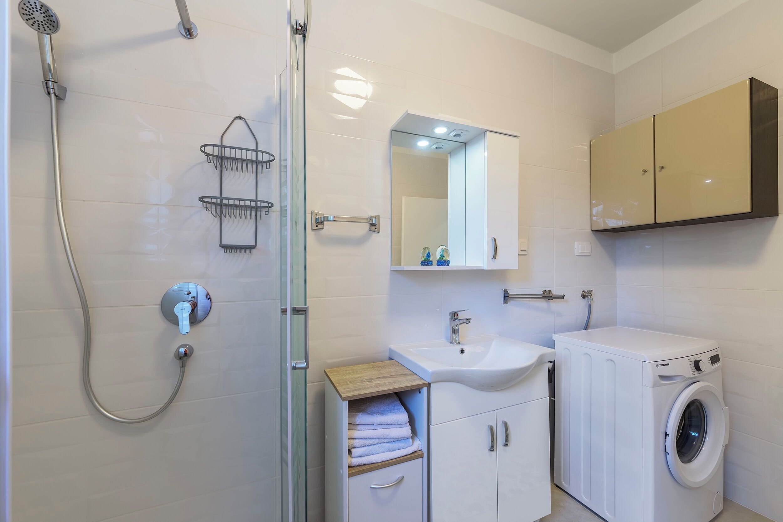 A modern bathroom with a glass-enclosed shower, a sink with a mirror above, and a white washing machine.