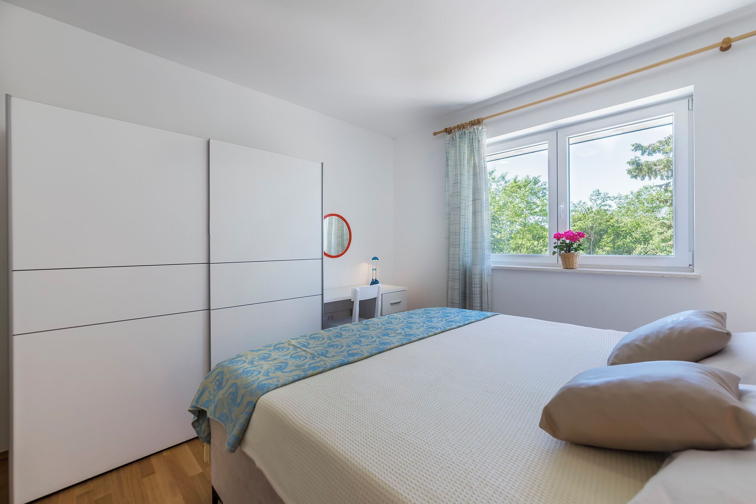Un dormitorio con cama, mesita de noche, espejo y ventana con vistas a árboles.