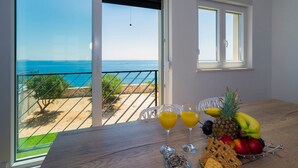 A balcony with a view of the sea, a table with fruit and drinks, and a window with a view of the beach. - Villa Adio Mare (Rogoznica)