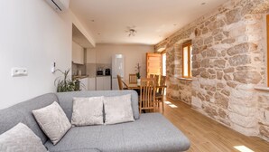 A modern living room with a grey sofa, a stone wall, and a kitchen in the background. - Villa Grgo (Rogoznica)