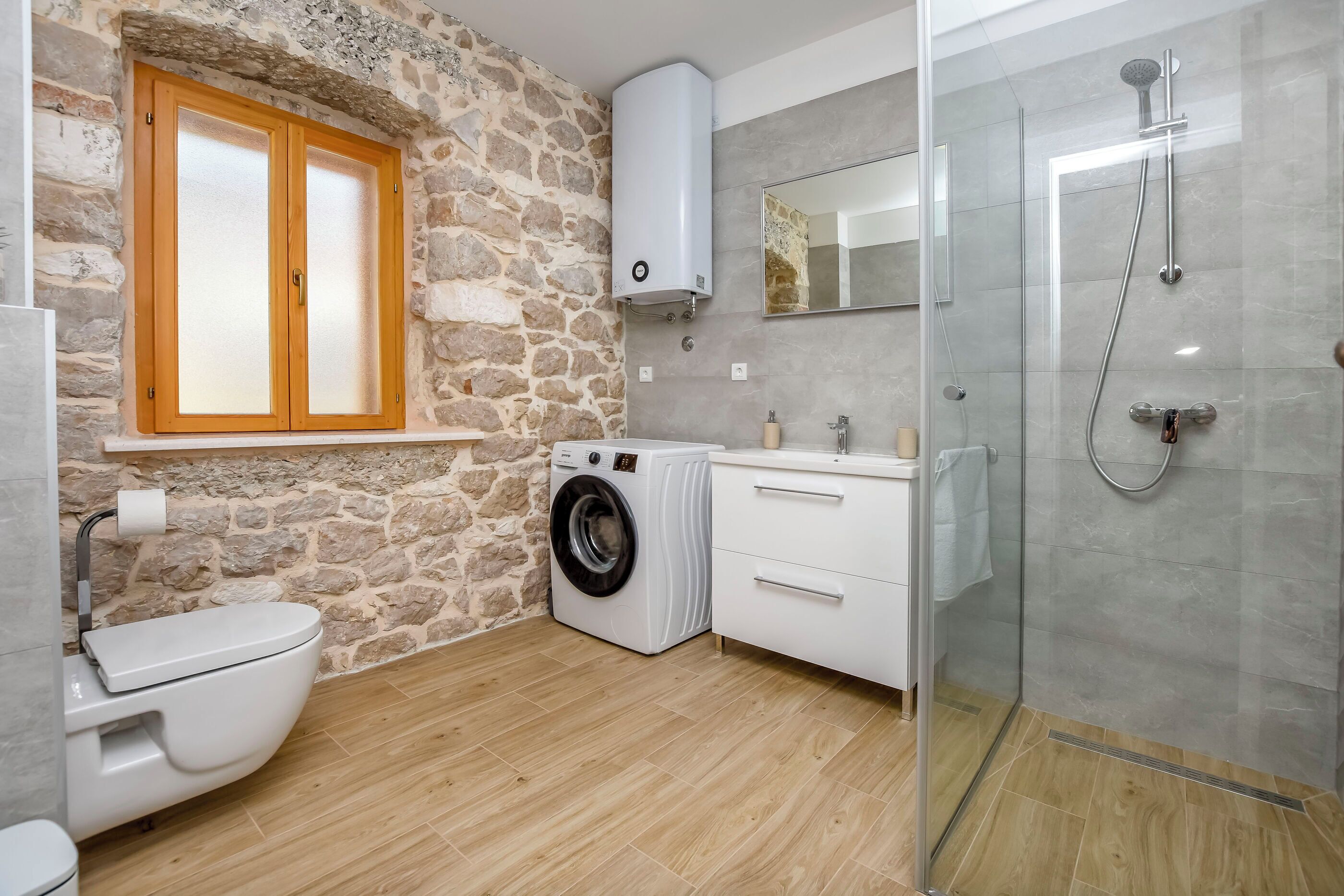 A modern bathroom with a stone wall, a washing machine, a toilet, and a glass-enclosed shower.