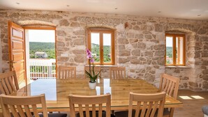 A dining area with a stone wall, wooden chairs, a glass table, and a view of the countryside through open doors. - Villa Grgo (Rogoznica)