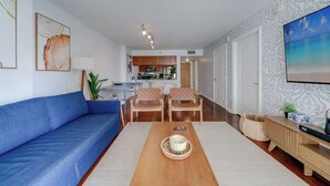 Una sala moderna con un sofá azul, una mesa de centro de madera y sillas de mimbre. Se ve una cocina al fondo con armarios de madera y un microondas.