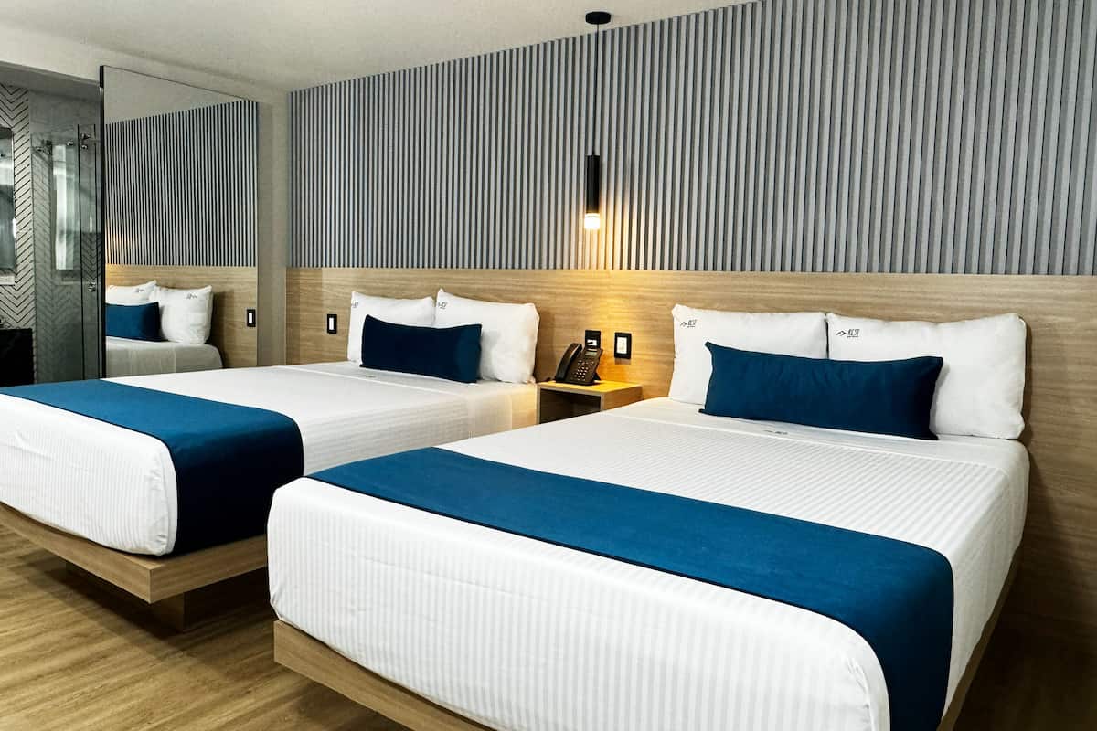 A hotel room with two beds, each with white bedding and blue accents, a wooden headboard, a bedside table with a lamp, and a striped wall.