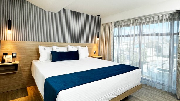 Ein modernes Hotelzimmer mit einem großen Bett, einem Holzkopfende und Blick auf die Stadt durch transparente Vorhänge.