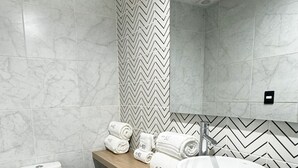 Ein modernes Badezimmer mit Toilette, Waschbecken und einem Spiegel darüber. Handtücher liegen auf einem Regal und der Boden ist mit schwarzen Fliesen ausgelegt.