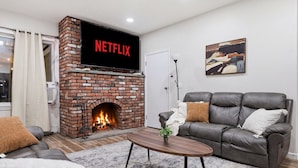 Una sala de estar con chimenea de ladrillo, un sofá de cuero gris, una mesa de centro y un televisor de pantalla plana mostrando 'NETFLIX'.