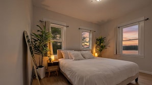 Un dormitorio con una cama grande, dos ventanas con cortinas, mesitas de noche con lámparas y plantas en macetas.