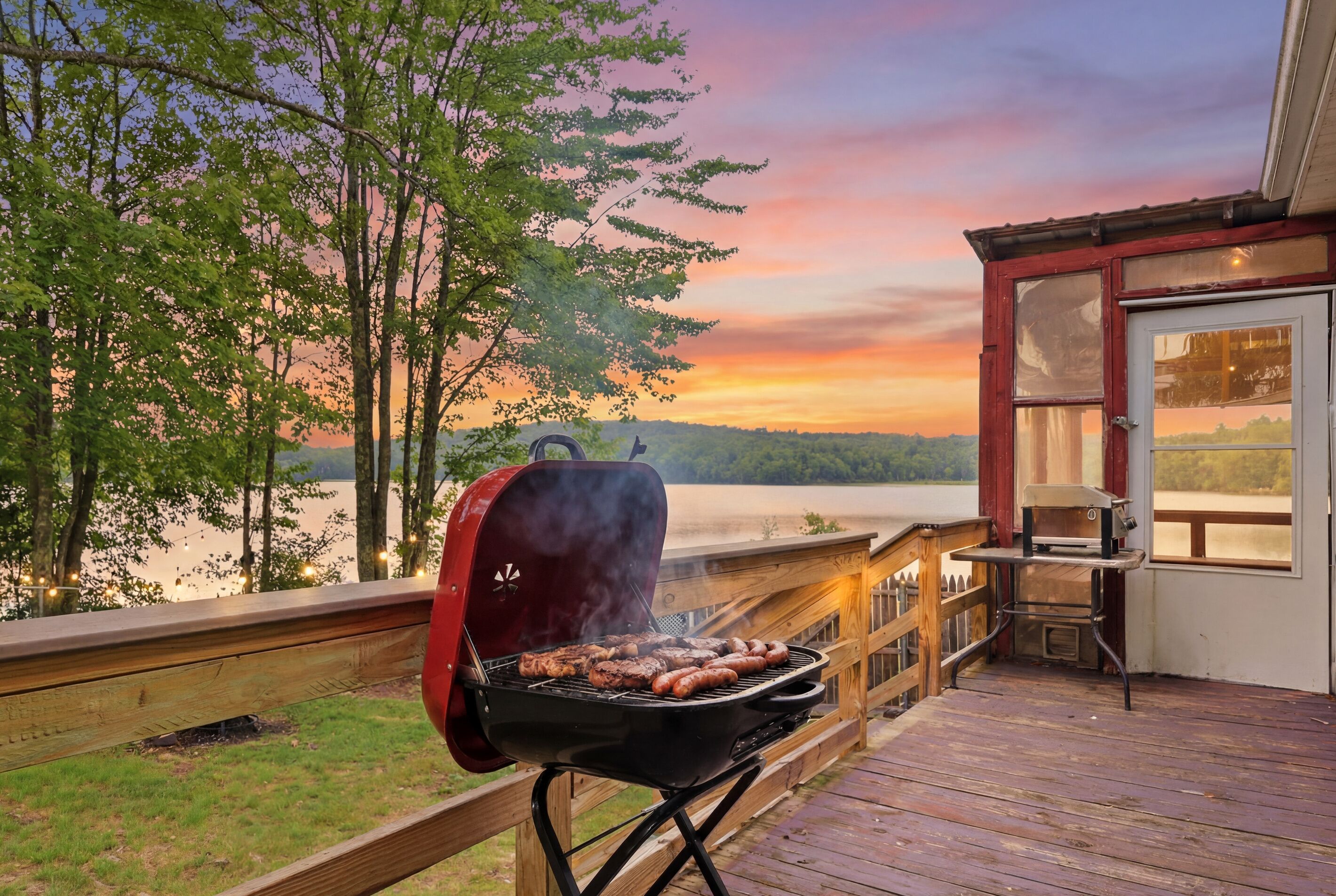 Una terraza de madera con una parrilla, barandilla de madera y vista al lago al atardecer.