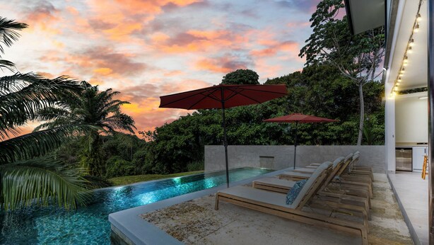 Ein moderner Poolbereich mit Liegestühlen und Sonnenschirmen, umgeben von üppiger Vegetation und einem Sonnenuntergangshimmel.