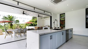Eine moderne Küche mit Marmorarbeitsplatte, hellblauen Schränken und einer großen Kochinsel. Es gibt eine Essecke mit Korbstühlen und einem Tisch sowie eine Terrasse mit Lichterketten und Sonnenschirmen.