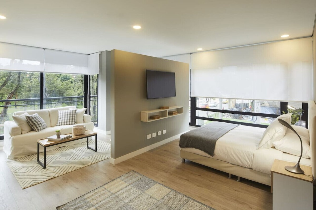 A modern bedroom with a large bed, a sofa, a TV mounted on the wall, and a balcony with a view of trees.