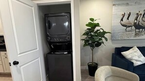 A compact living space with a kitchenette, a seating area with a sofa and armchair, a potted plant, and a wall-mounted artwork of pelicans. - Kapilana - Pelican Unit H- Lakefront w/Boat Rental (Osage Beach)