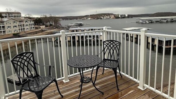 A wooden deck with two metal chairs and a table, overlooking a marina with boats and a distant shoreline. - Kapilana - Pelican Unit H- Lakefront w/Boat Rental (Osage Beach)