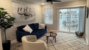 A living room with a blue sofa, a white armchair, a round coffee table, and a painting of birds on the wall. - Kapilana - Pelican Unit H- Lakefront w/Boat Rental (Osage Beach)
