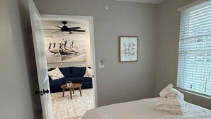 A bedroom with a bed, a ceiling fan, a window with blinds, and a door leading to a living area with a sofa and a table. - Kapilana - Pelican Unit H- Lakefront w/Boat Rental (Osage Beach)