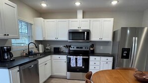 A modern kitchen with white cabinets, stainless steel appliances, a wooden dining table, and a window with blinds. - Kapilana Beach Unit I - Lakefront! w/Boat rental (Osage Beach)