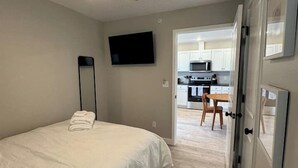 A bedroom with a bed, a ceiling fan, a TV, and a view into a kitchen area. - Kapilana Beach Unit I - Lakefront! w/Boat rental (Osage Beach)