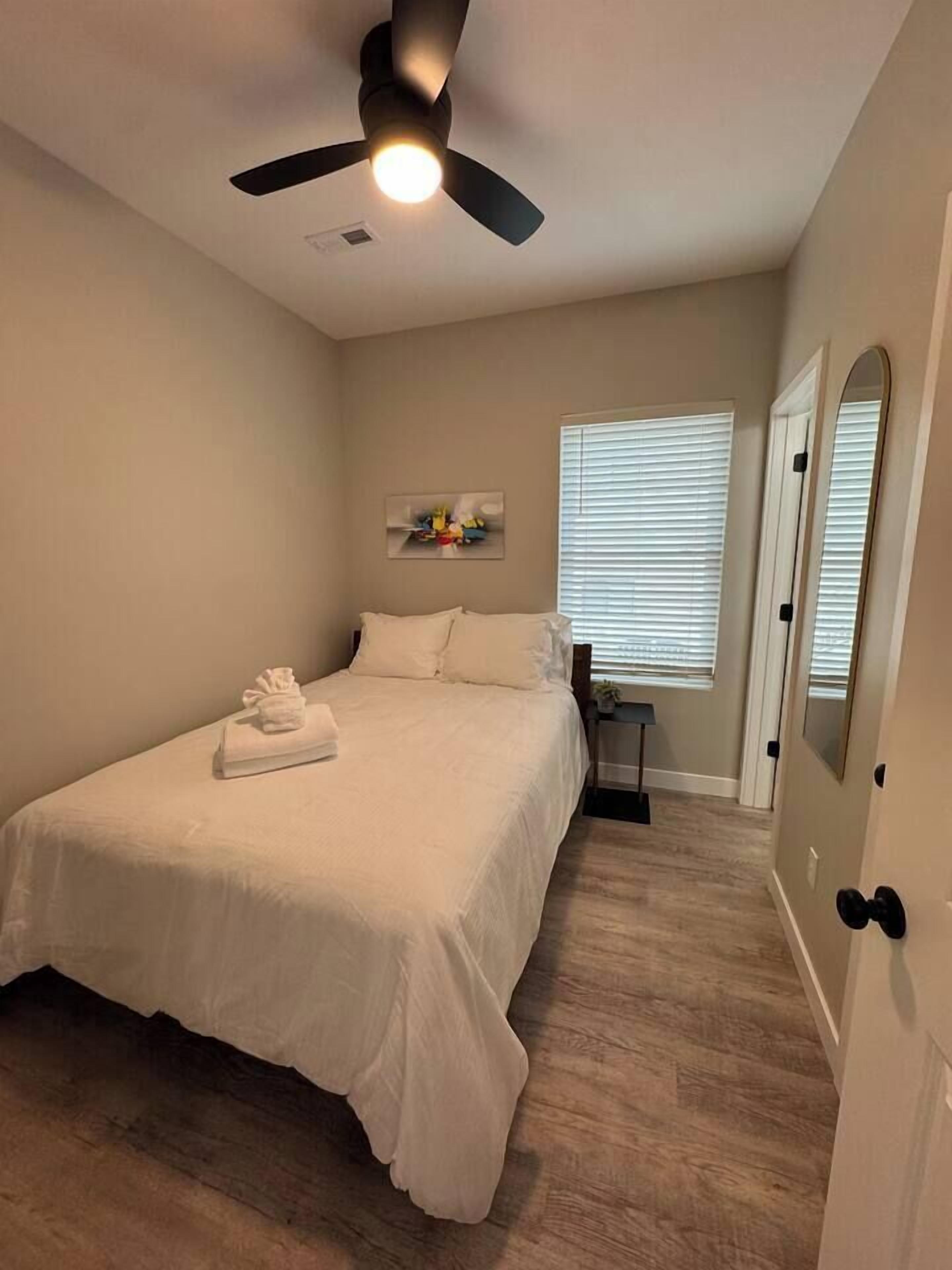 A bedroom with a bed, a ceiling fan, a window with blinds, and a door with a mirror.