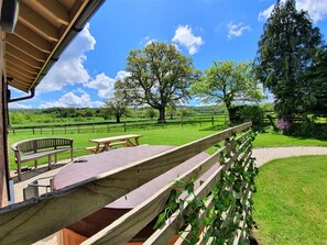 Terraza de madera con mesas de picnic y bancos, con vistas a un paisaje verde con árboles y cielo despejado.