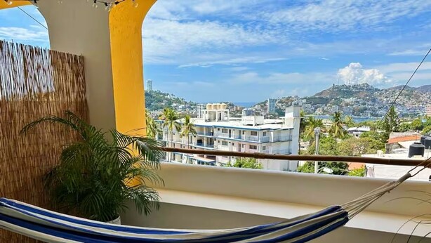 A balcony with a hammock, a potted plant, and a view of a cityscape with buildings and hills. - Loft with ocean view! "Twin Towers Tres Mares" (Acapulco de Juárez)