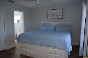 A bedroom with a bed, drawers, a ceiling fan, and a framed sign on the wall.