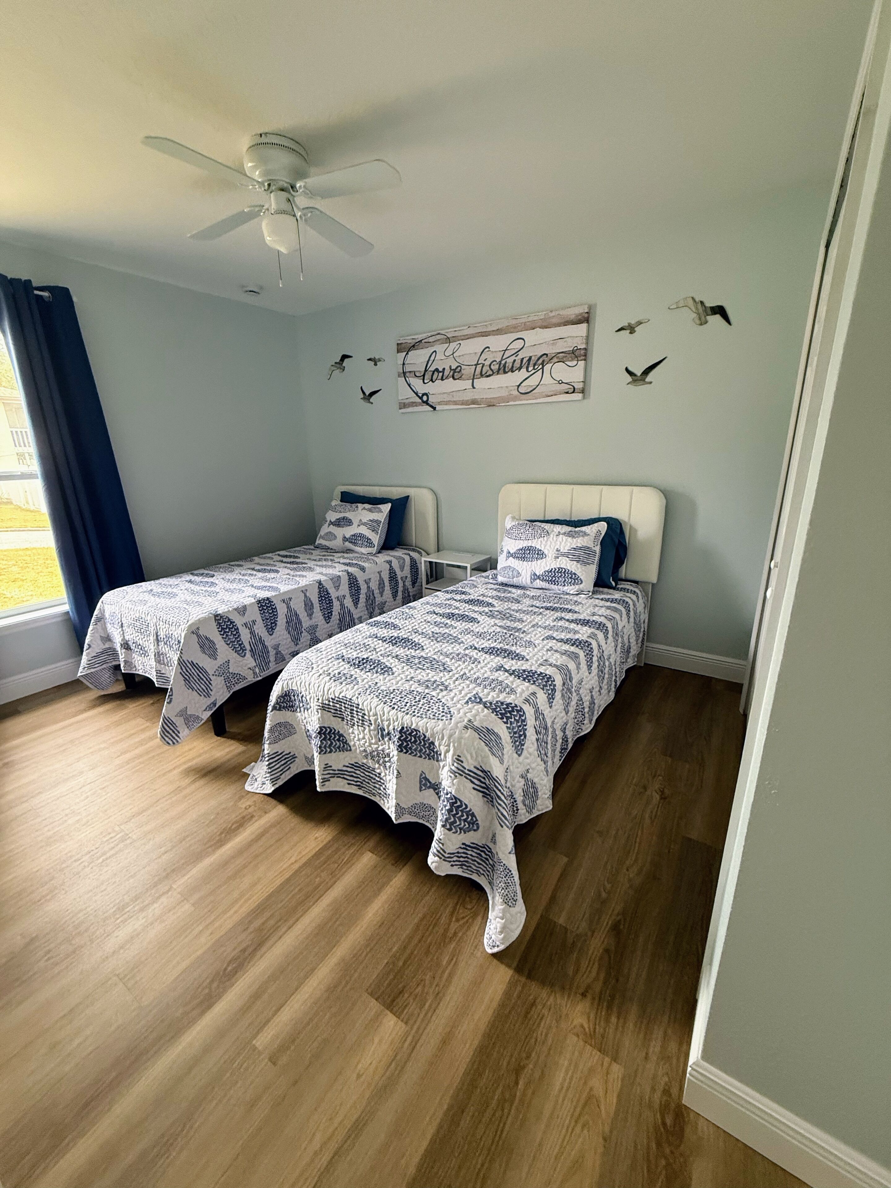 A bedroom with two beds, a ceiling fan, a wall decoration with birds and the text "Love fishing," and a window with blue curtains.