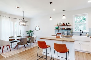 A modern kitchen with a central island, bar stools, a dining area with chairs, pendant lights, and a view of the outdoors through a window. - Savannah Retreat | Private Spa | 6 min to Downtown (Savannah)