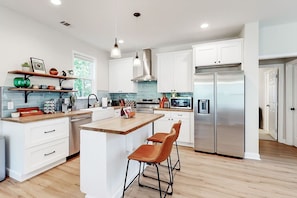 A modern kitchen with white cabinetry, stainless steel appliances, a central island with bar stools, and a window providing natural light. - Savannah Retreat | Private Spa | 6 min to Downtown (Savannah)