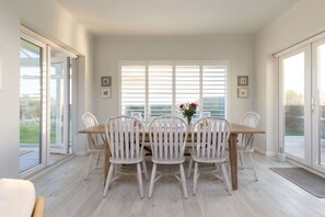 A dining area with a wooden table and white chairs, large windows with blinds, and a vase with flowers on the table. - Luxury Beachfront Retreat for 14 With Hot Tub (Bognor Regis)