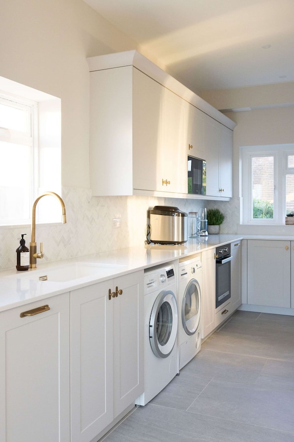 A modern kitchen with white cabinetry, a built-in washing machine, and a window providing natural light. - Luxury Beachfront Retreat for 14 With Hot Tub (Bognor Regis)
