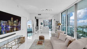 A modern living room with a large TV, a sofa, a coffee table, and a view of a cityscape through large windows. - Luxury Corner Unit with 3 Private Balconies and Ocean Views (Miami)