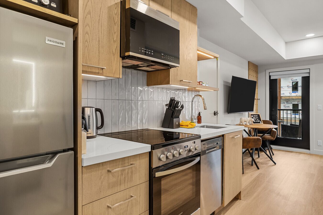 A modern kitchen with a stainless steel refrigerator, a built-in oven, and a stove. There is a sink with a faucet, a countertop, and a chair.