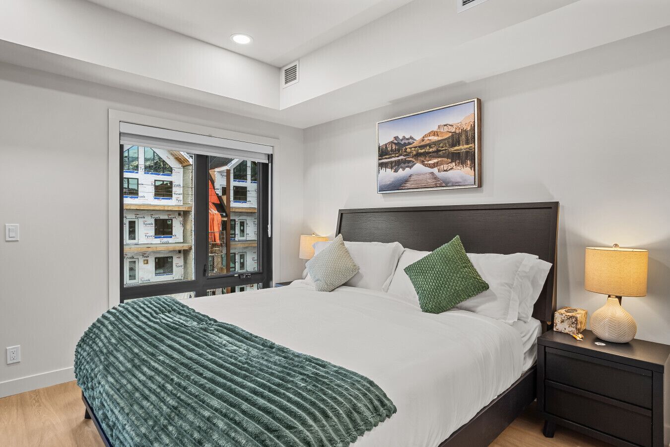 Un dormitorio con cama, mesita de noche, lámpara, ventana con vistas a edificios y un cuadro en la pared.