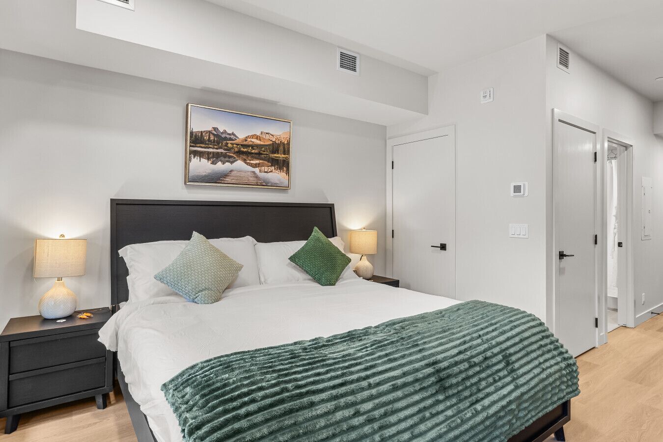 A modern bedroom with a large bed, two bedside tables with lamps, a painting above the bed, and a door to the right.