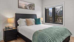 A bedroom with a bed, two bedside tables, a window with a mountain view, and a framed picture above the bed. - “The Ascent Mountain Haven” (Canmore)