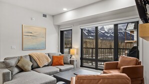 A modern living room with a grey sectional sofa, a brown leather armchair, and a coffee table. There is a large window with a view of mountains and a balcony with a railing. - “The Ascent Mountain Haven” (Canmore)