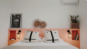 Una cama bien tendida con sábanas blancas y dos almohadas a rayas negras, un aire acondicionado de pared, un cuadro enmarco, un adorno de pared y una planta en maceta.
