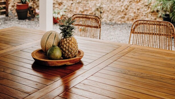 Una mesa de madera con una lámpara colgante de mimbre, rodeada de sillas de mimbre, en un espacio exterior cubierto con plantas y una estructura blanca al fondo.