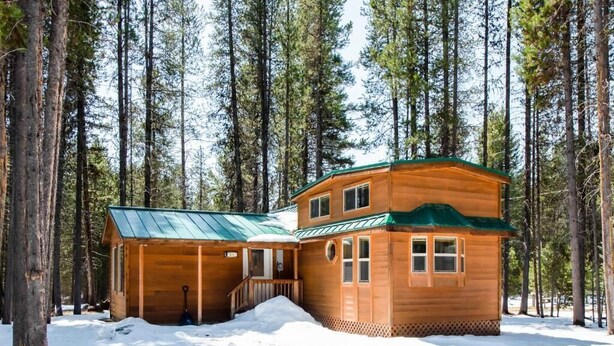 Una cabaña de madera con techo verde, rodeada de altos pinos cubiertos de nieve.