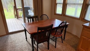 Un comedor con una mesa de madera y cuatro sillas, suelo alfombrado y ventanas amplias con persianas.