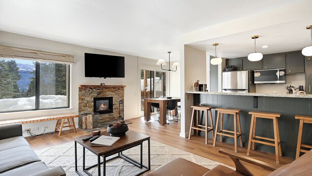 A modern living room with a fireplace, a large window, and a kitchen area with bar stools. - Discovery 4 111:Near Canyon Lodge, Dog Friendly (Mammoth Lakes)