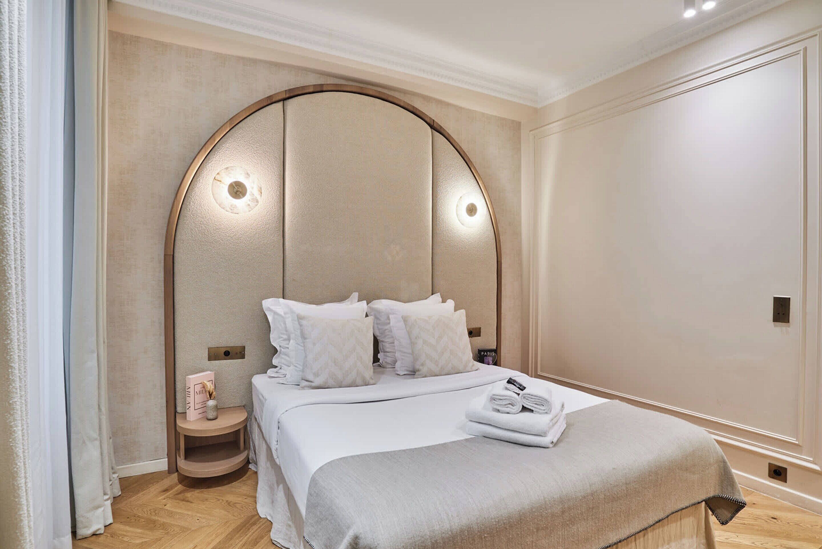 Habitación de hotel con una cama grande, una mesita de noche de madera y un cabecero curvo con luces.