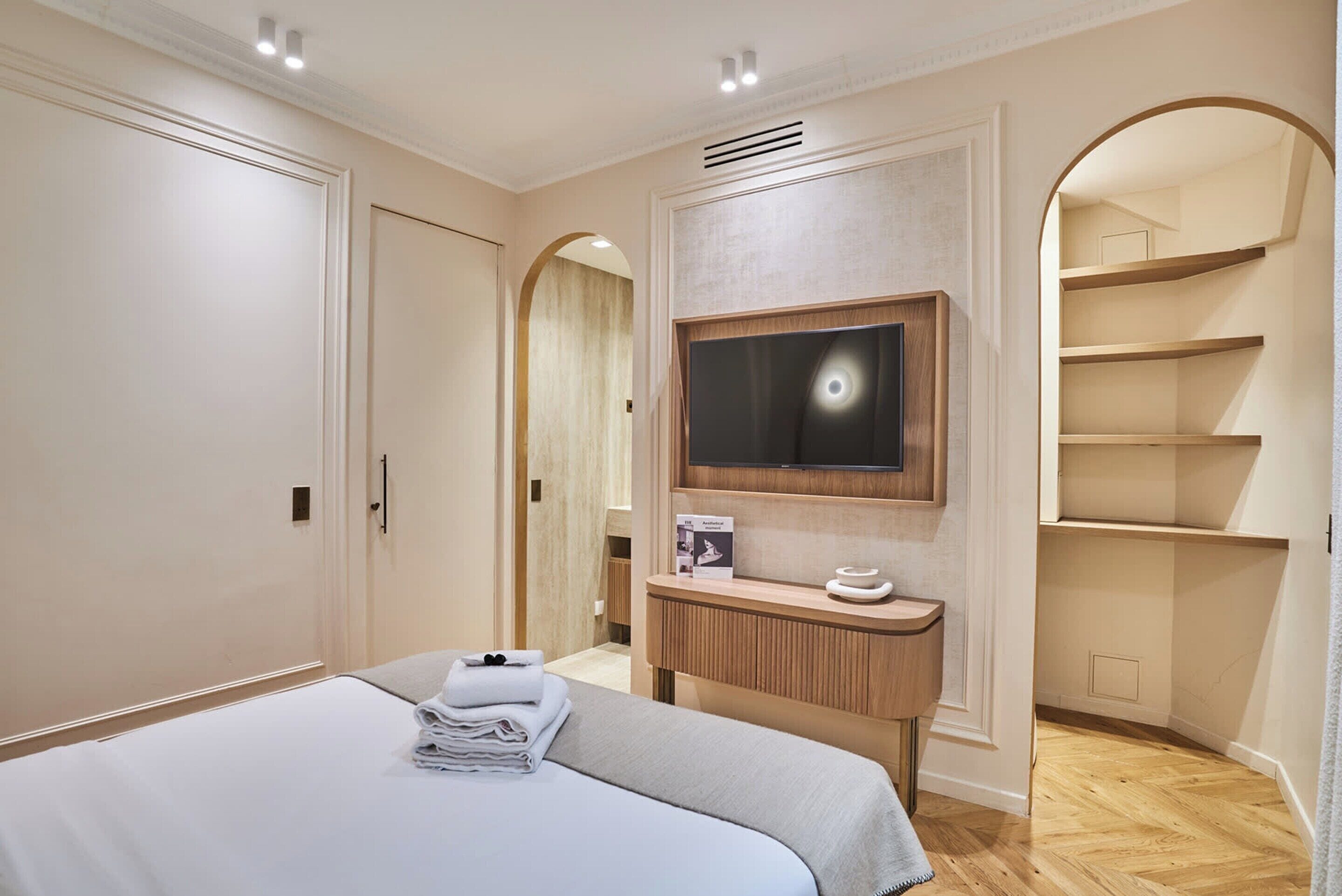 A hotel room with a bed, a TV mounted on the wall, a vanity with a mirror, and built-in shelving.