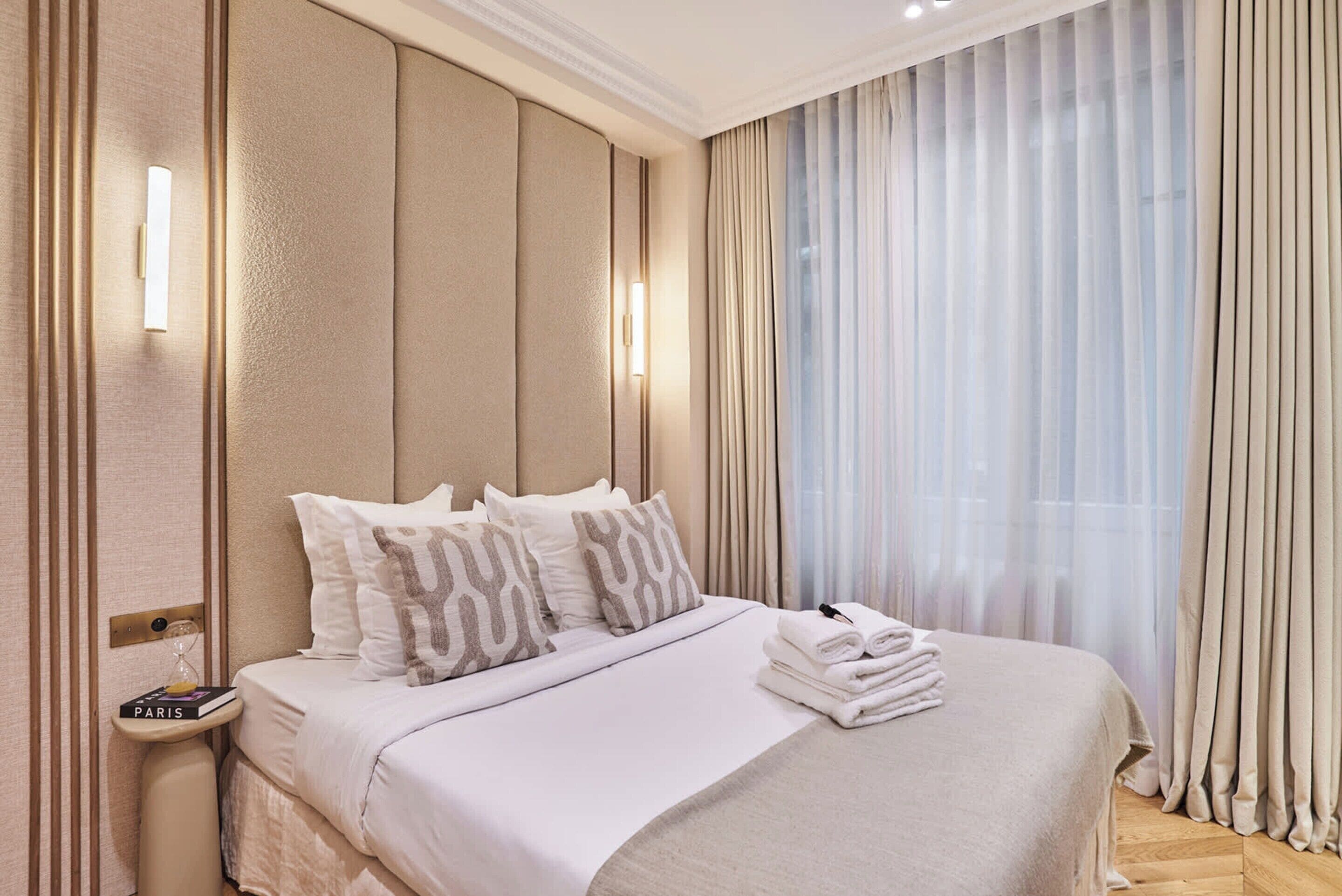 Habitación de hotel con una cama grande, una mesita de noche con lámpara y cortinas translúcidas en la ventana.
